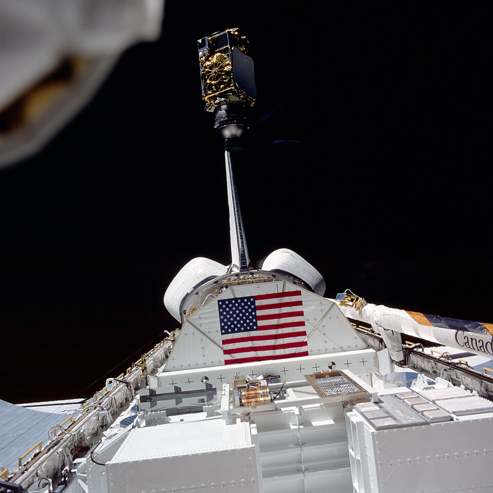 The INSAT-1B satellite on its PAM-D booster lifting clear of the Space Shuttle payload bay against black space, the Canadarm and a US flag visible below