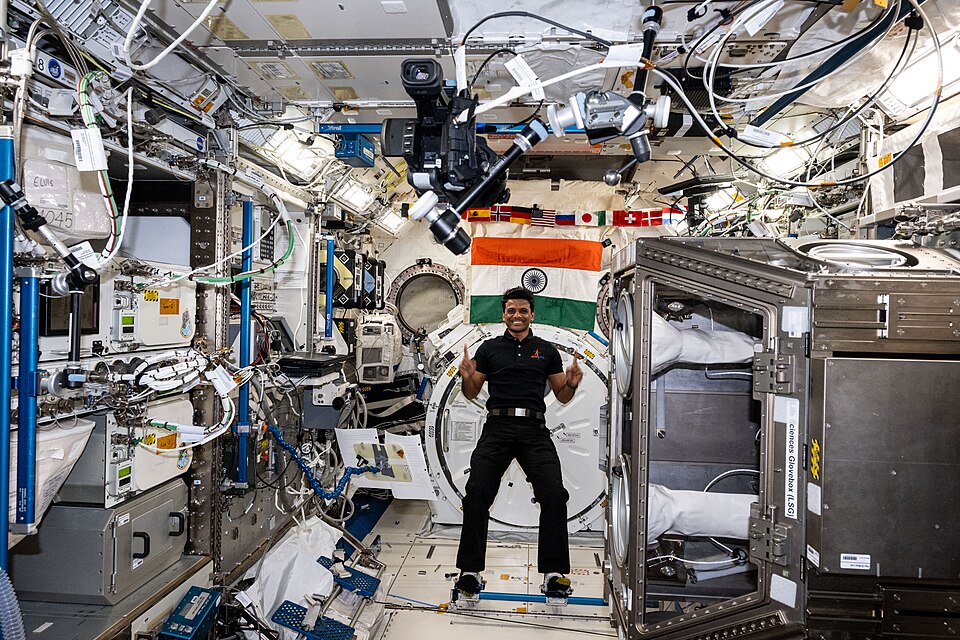 Astronaut Shubhanshu Shukla floating in a module of the International Space Station, both hands raised, with the Indian tricolour mounted on the wall behind him