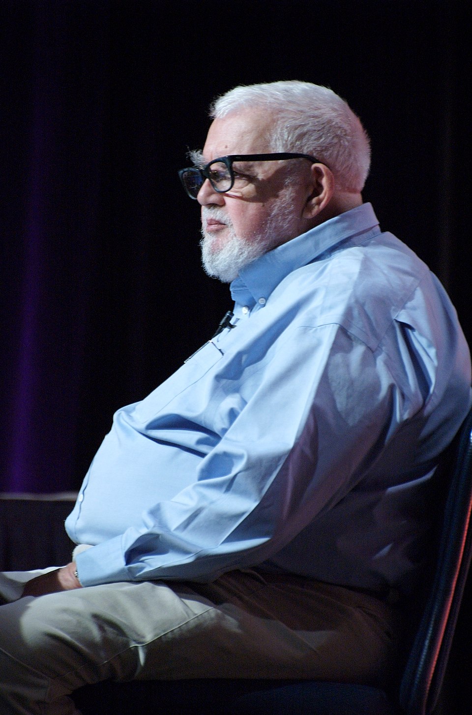 Side-on portrait of John McCarthy, white-haired and white-bearded in heavy black spectacles and a pale blue shirt, seated on a darkened stage