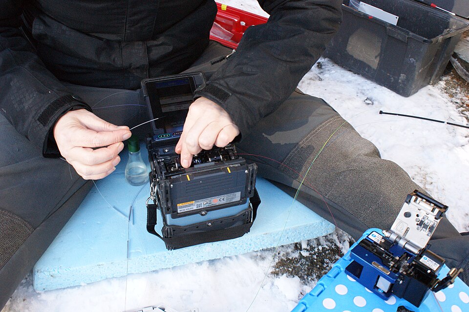 A technician feeding a bare optical fibre into an open fusion splicer resting on a foam board, with a fibre cleaver beside it