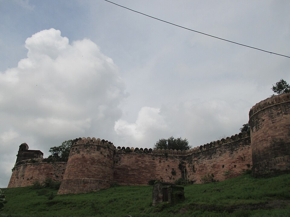 The red sandstone ramparts and round bastions of Dhar Fort rising above a grassy slope under monsoon clouds