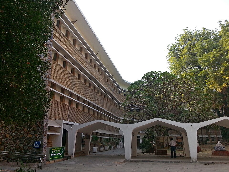 The modernist Rabindra Bhavan in New Delhi, a long brick-and-concrete block with ribbon balconies, seen from its shaded forecourt with a bookshop sign at the entrance