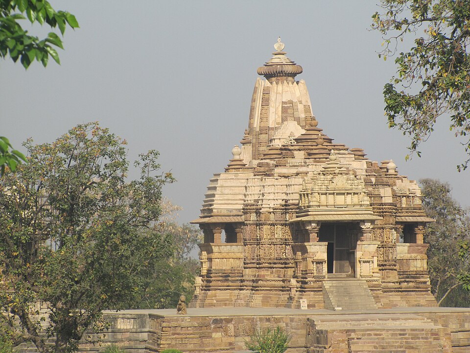 The sandstone Devi Jagadambi Temple at Khajuraho, its tiered shikhara tower rising above a carved body and pillared entrance porch, framed by trees