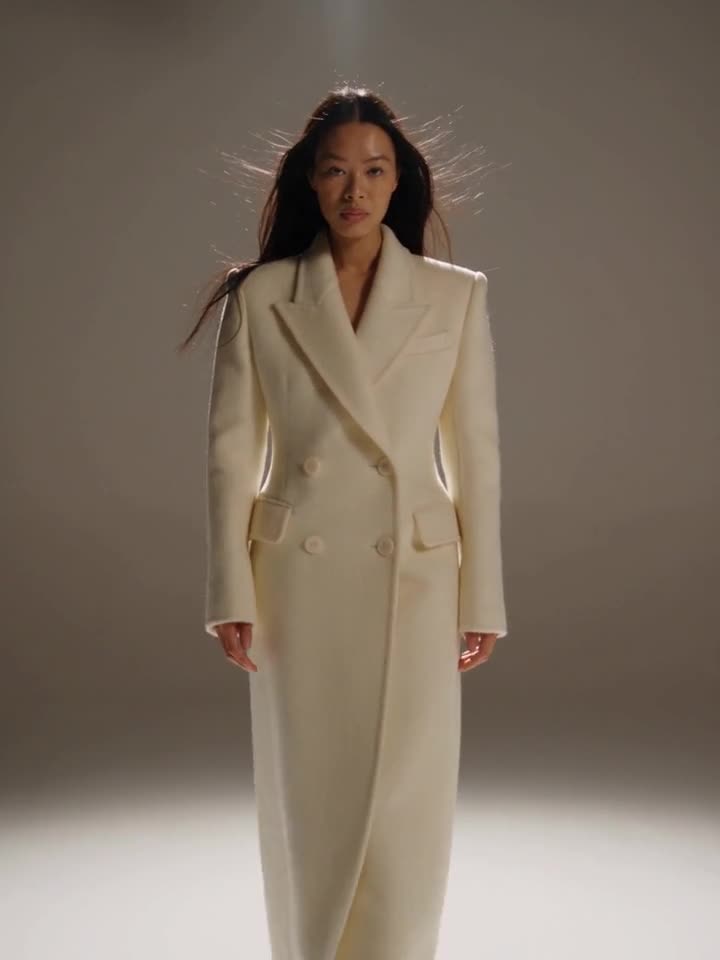 Alexandra Lim, in a long ivory coat walking toward camera in a grey studio