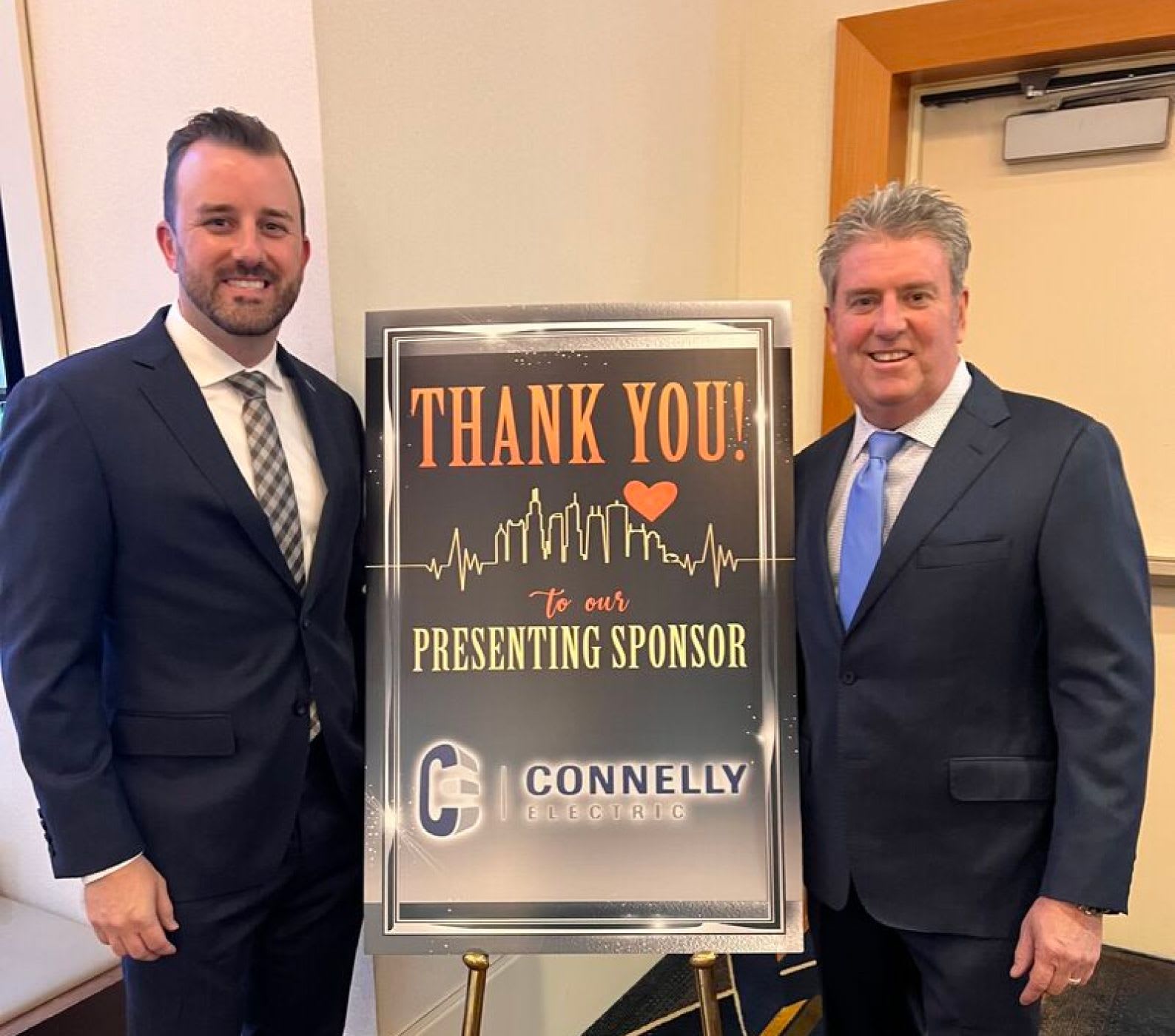 Two Connelly leaders dressed in suits and standing next to a sign that says "Thank you to our presenting sponsor, Connelly Electric". 
