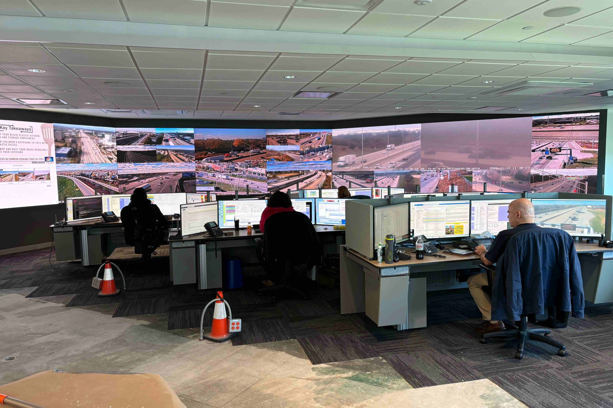 Three people working at their desk in front of a video wall showing Illinois roadways.