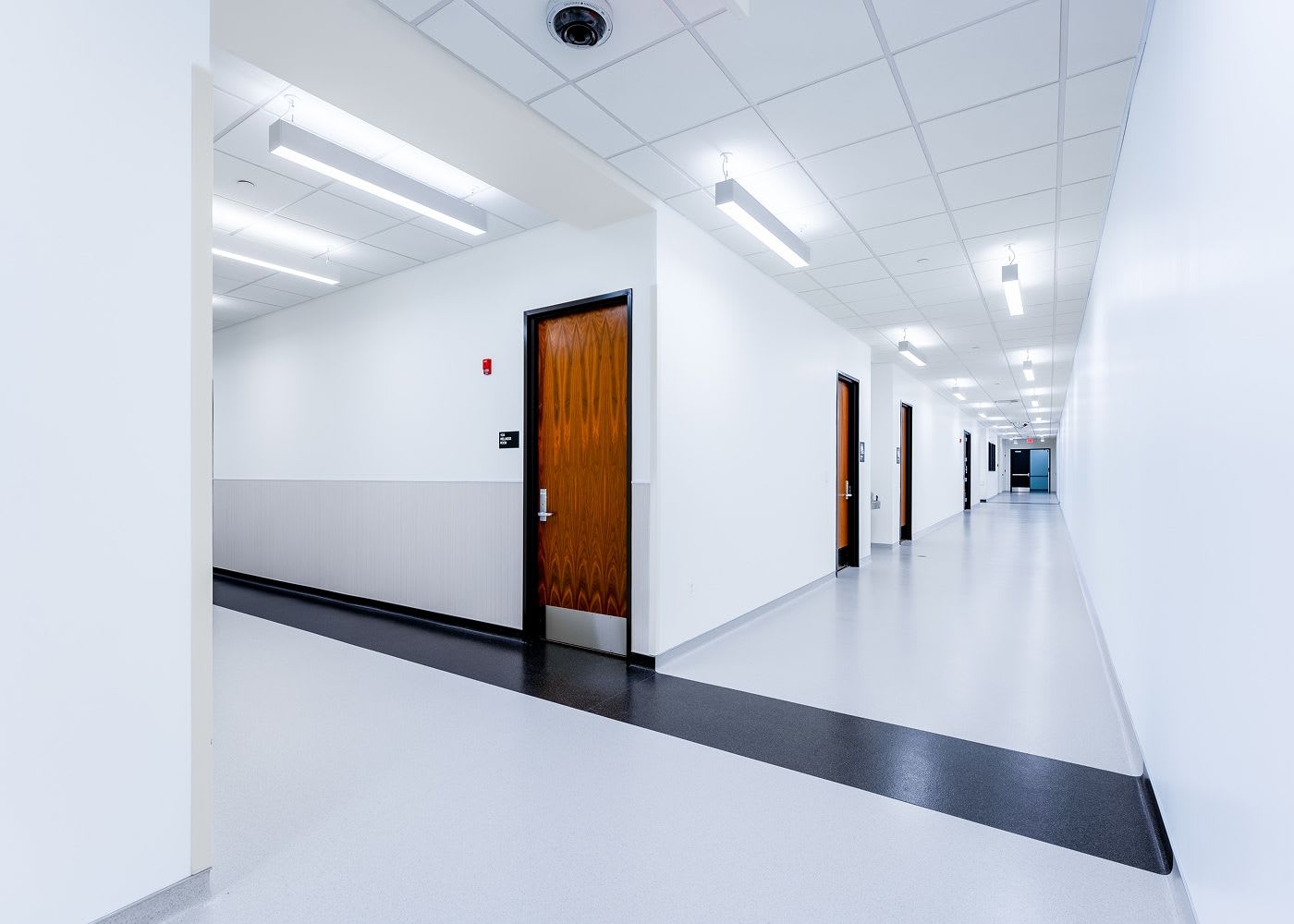 A long and bright lit hallway with white walls and several closed wood doors.