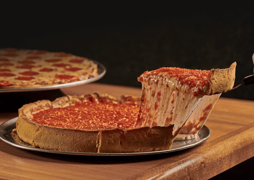 A steamy slice of deep-dish pizza from Beggars Pizza is lifted with a pie server. A pepperoni pizza sits in the background.