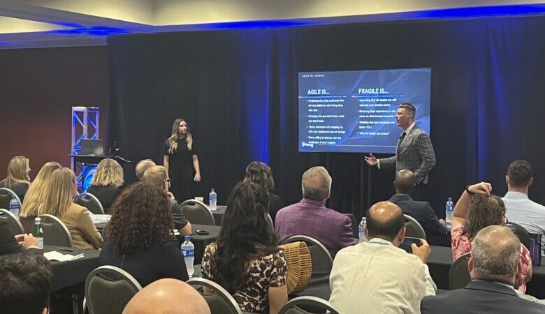 a room of people sitting looking at two people presenting in front of a large screen