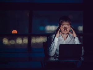 Distressed man holding his temples at a desk with his laptop open in a dark room 
