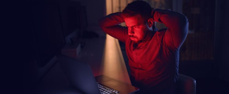 Stressed out man holding hands behind head, blowing out cheeks, and looking at a laptop