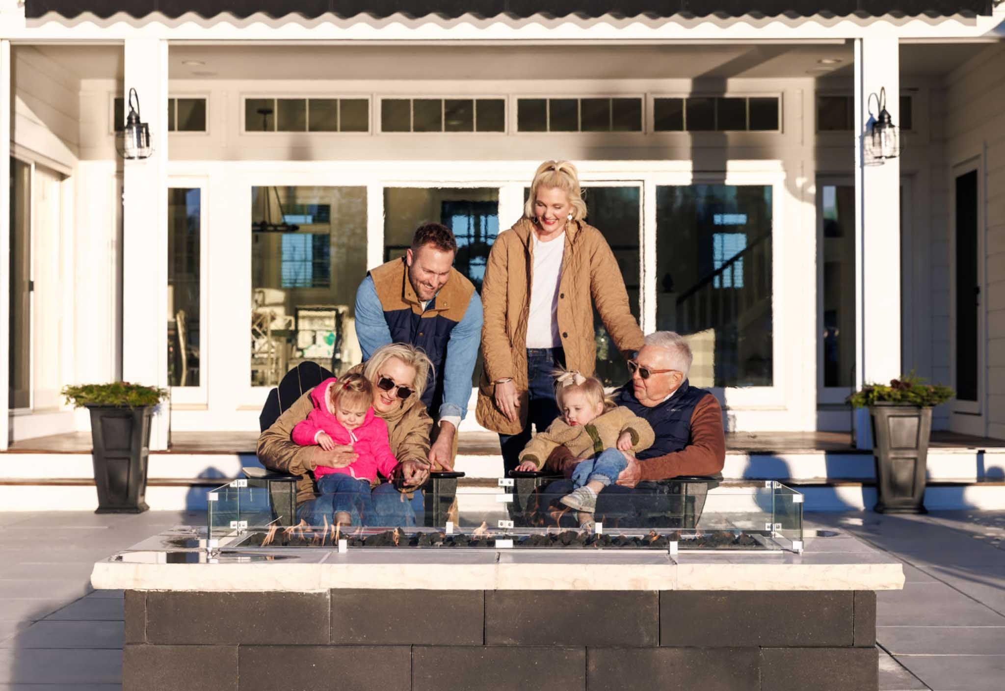 A mother, father, grandparents, and two young girls gather around an outdoor fire pit at their home in Geneva National Community