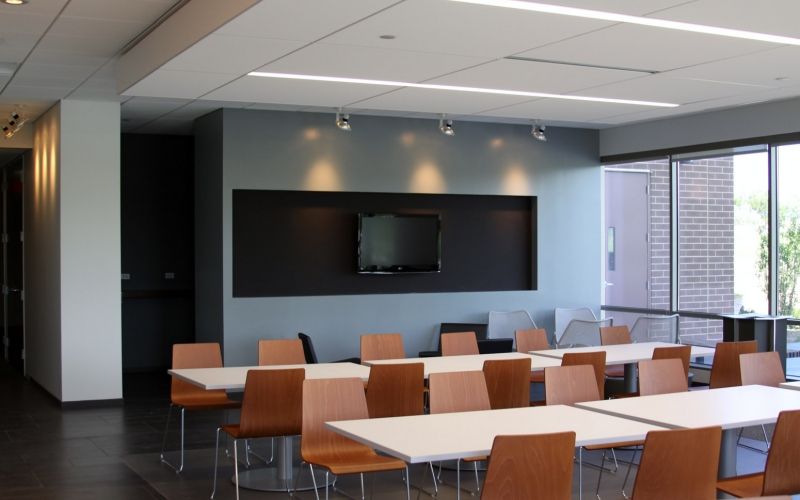 Dining room hall with a television, chairs, glass windows and five white tables.