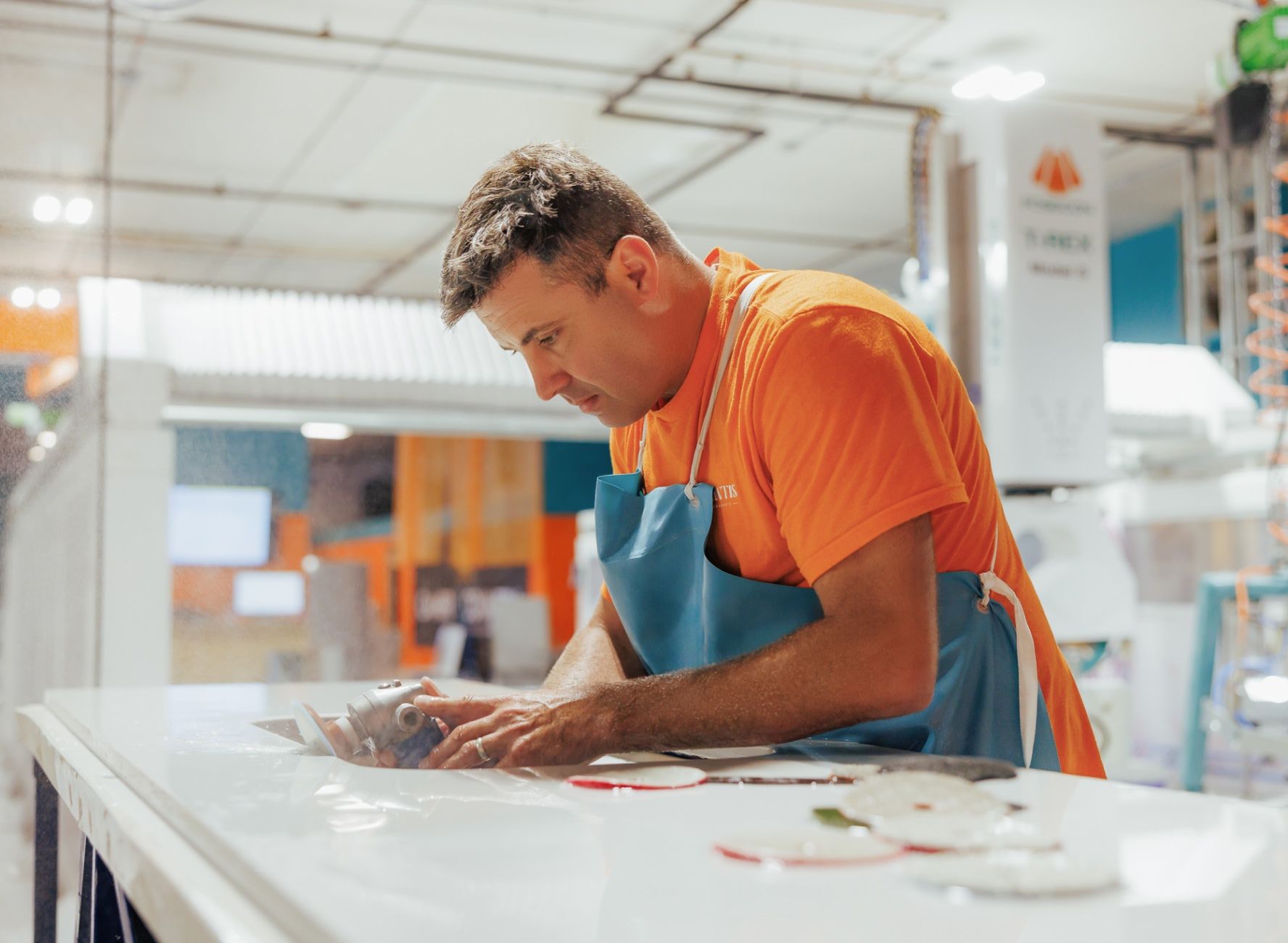 Fabrication Shop Worker sanding down countertop stone slab for sink faucets wearing an orange t shirt and teal apron