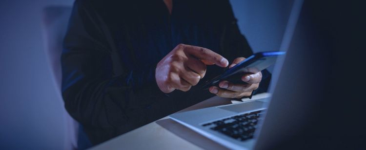 A person using a smartphone in front of a laptop - likely authenticating their login with MFA