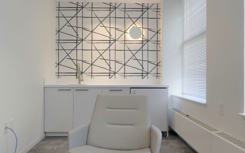 Small, white room with a chair, some cabinets, a sink and a decorative line wallpaper.