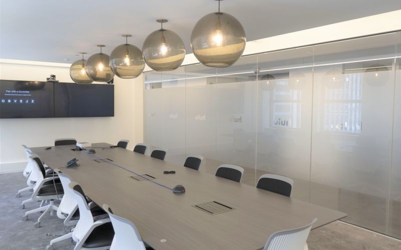 Meeting room with a long wooden table, screens, glass walls and rounded light fixtures.