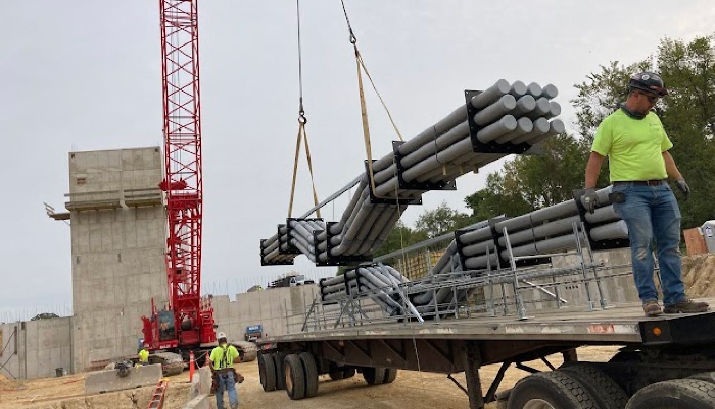 A crane lifting prefabricated electrical multi-conduit racks at a construction site.
