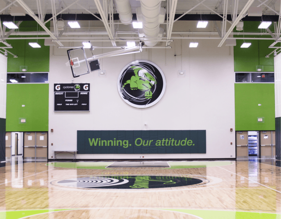 An indoor basketball court with wood floors, large ceiling lights and a sign on the wall that says "Winning. Our attitude".