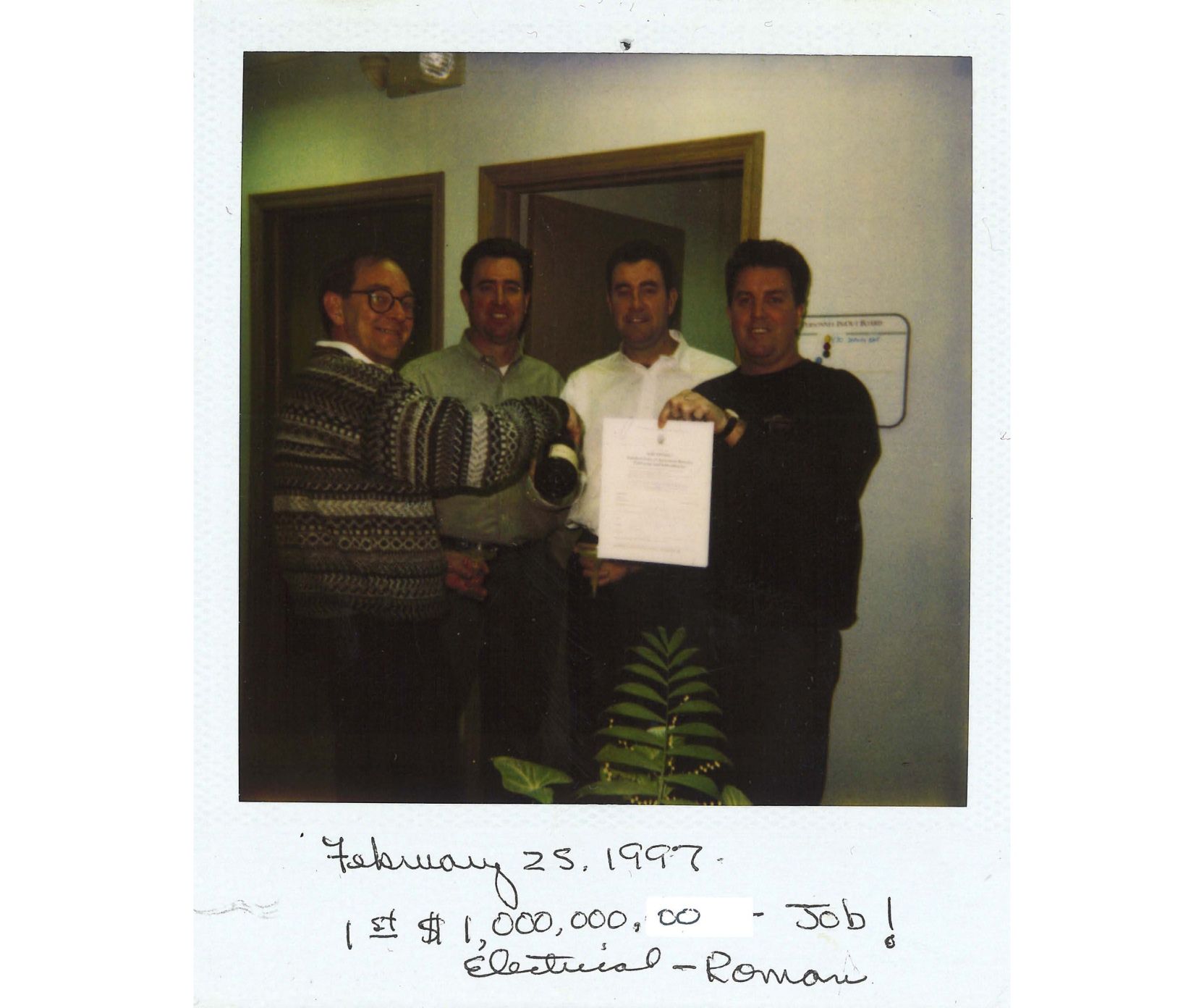 Four Connelly members standing next to each other, smiling and holding a piece of paper with a caption below the photo stating "February 25, 1997. 1st $1,000,000.00 Job!"