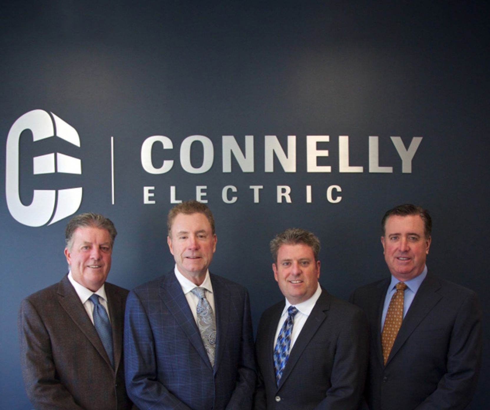 Four Connelly leaders wearing suits and standing next to each other smiling in front of a wall with the Connelly Electric logo.