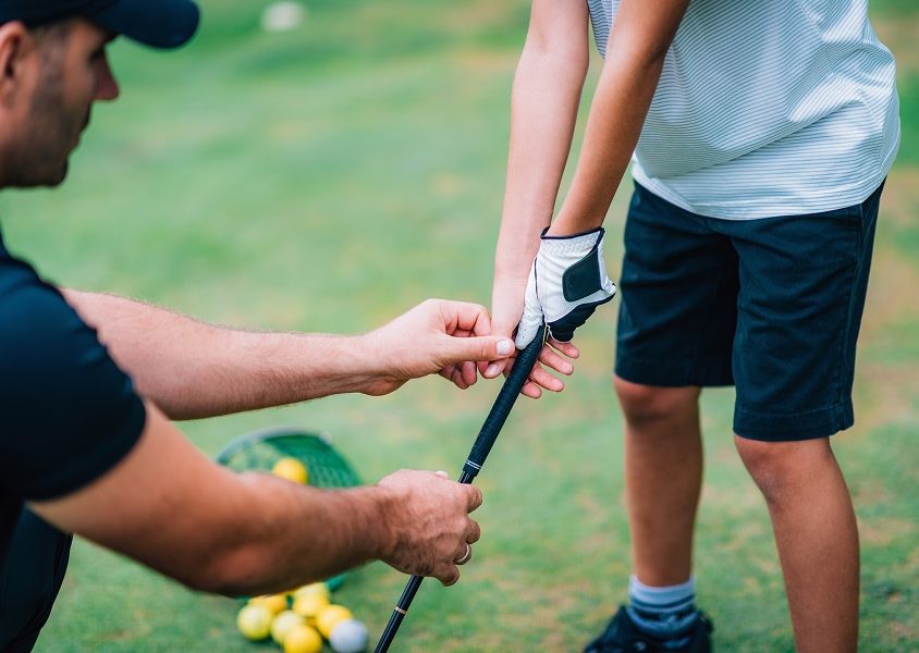 A golfer helps a young golfer adjust his grip on the club