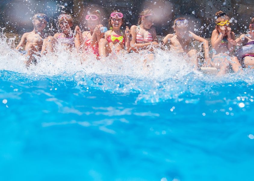A line of kids sit at the edge of a pool splashing the water with their feet