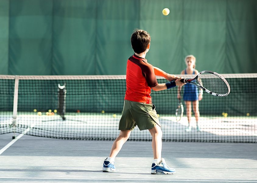 A young boy stands on the tennis court with his back to the camera, having just hit the ball in the direction of his opponent, a young girl standing opposite him