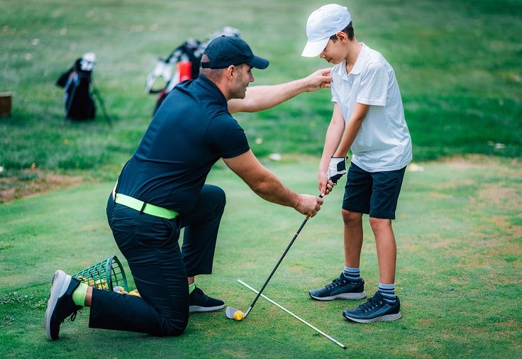 A golfer kneels to help a younger golfer adjust his form as he prepares to hit the ball