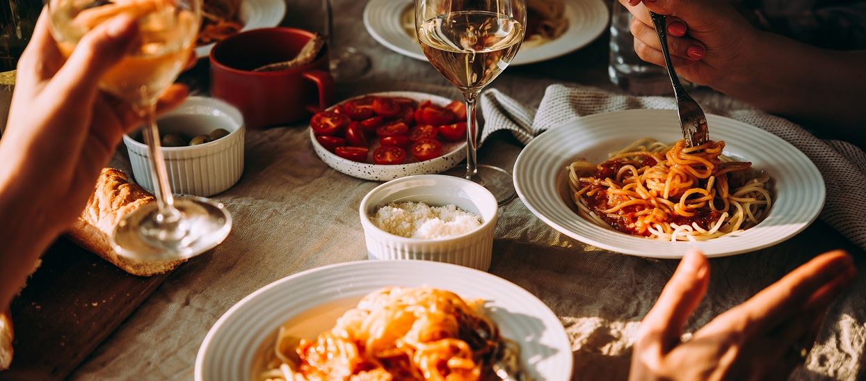 Hands interact with plates of pasta, tomatoes, and parmesan as well as wine glasses