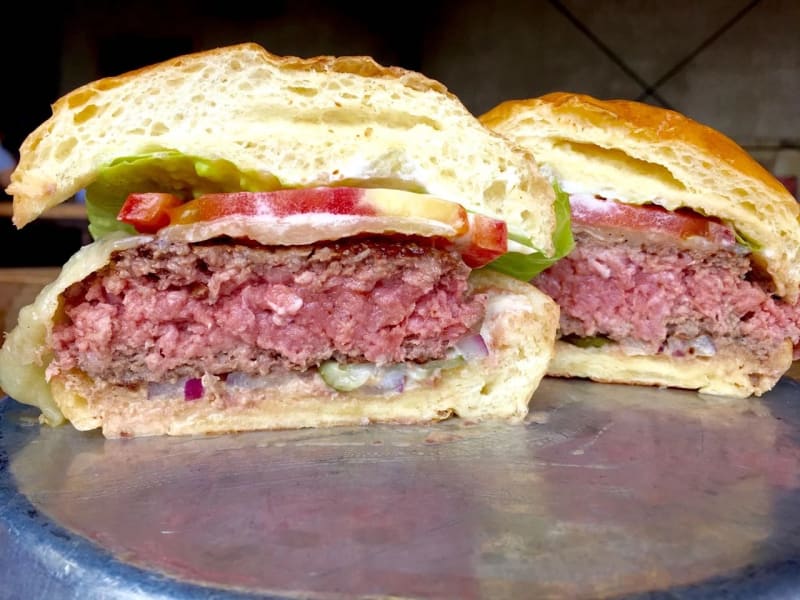 The Contigo burger cut in half on a metal tray: a rare patty on house challah with tomato, lettuce, red onion, and pickles