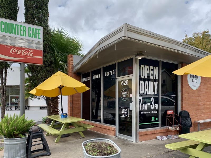 The original Counter Cafe at 626 North Lamar: a small brick storefront with Open Daily 7 AM to 3 PM on the window, yellow umbrellas, and green picnic tables out front