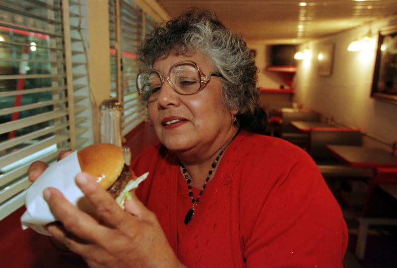 A woman in a red sweater and glasses holds up a Fran's burger in its paper wrapper at a window booth inside the diner