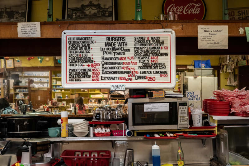 The Nau's Enfield Drug lunch counter: a lit menu board listing hamburgers, cheeseburgers, shakes, and malts, with a Coca-Cola sign above and the grill below