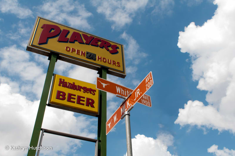The Player's sign against a cloudy blue sky, reading Open 24 Hours and Hamburgers Beer, beside the Whitis Avenue and W. MLK Jr. street signs