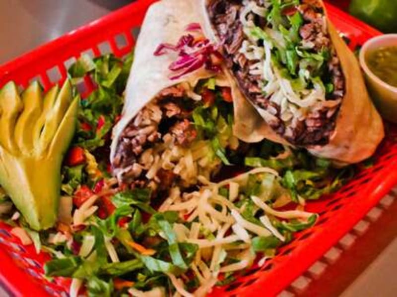 A Changos burrito cut in half in a red plastic basket, stuffed with steak, lettuce, cheese, and red cabbage, with sliced avocado and salsa verde on the side