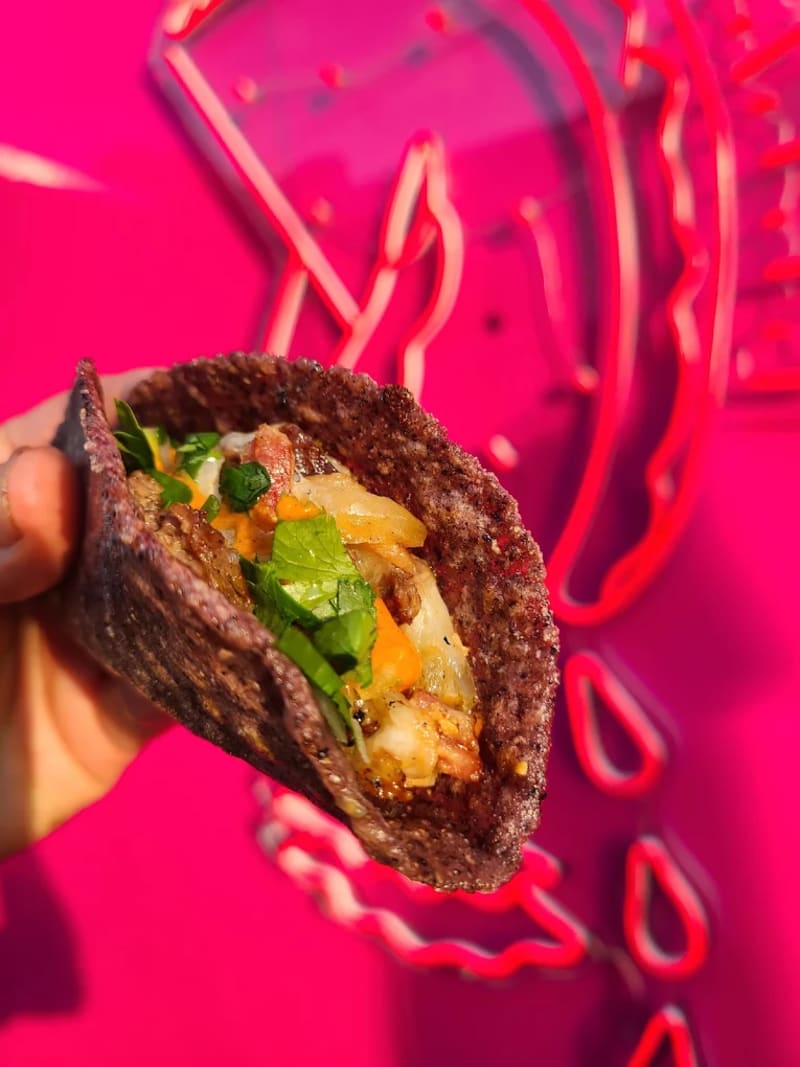 A Con Todo taco held up in one hand against a pink neon sign: chopped meat, onion, cilantro, and salsa on a blue corn tortilla