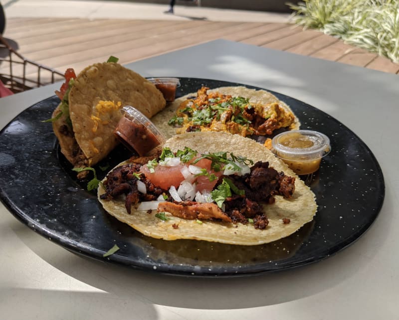 Three Dai Due Taqueria tacos on a black speckled plate on the Fareground patio: wild boar al pastor with grapefruit, onion, and cilantro in front, a picadillo taco and a crispy taco behind
