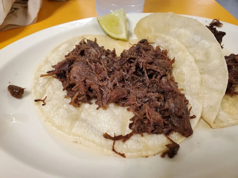 Two La Mexicana barbacoa tacos on a white plate: shredded barbacoa piled on corn tortillas with a lime wedge, at an orange counter