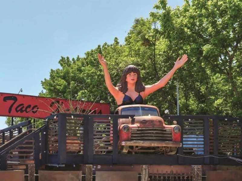 The giant bust of Maria on the roof of Taco Xpress on South Lamar, arms raised over a rusted vintage pickup, with the red Taco Xpress sign and trees behind