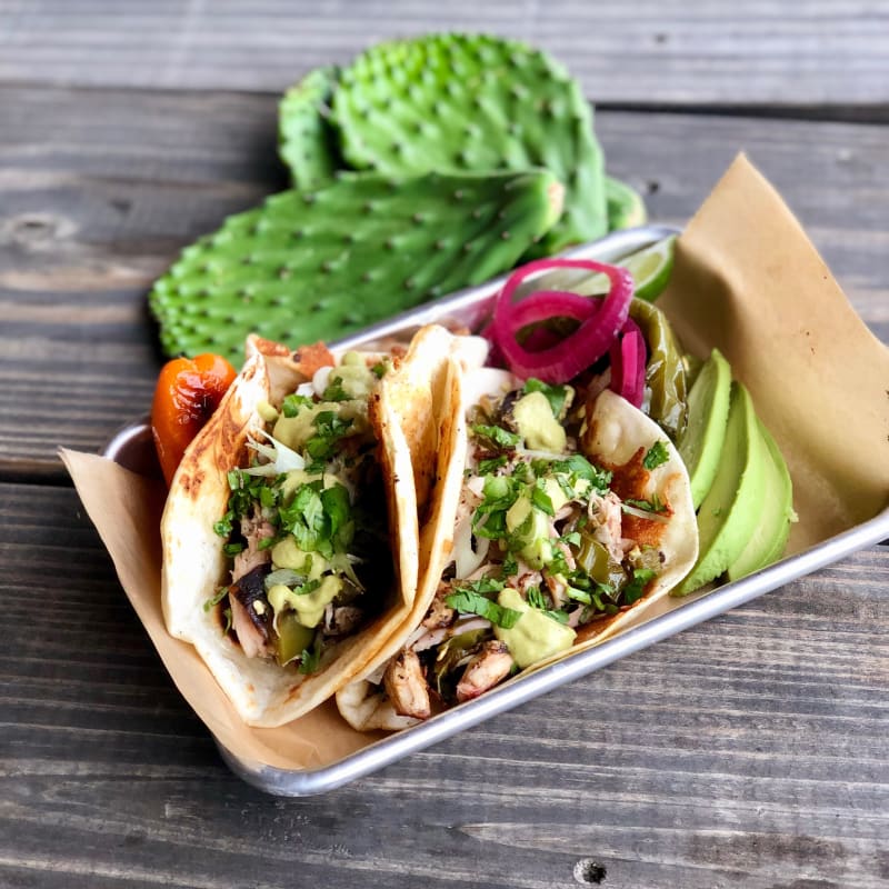 Two Taco Bronco tacos in a paper-lined metal tray: smoked meat with cilantro, onion, pickled red onion, and avocado, with nopal paddles on the picnic table behind