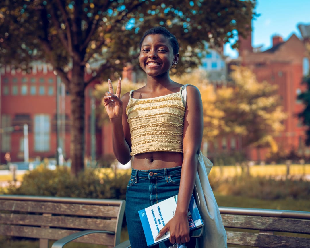 El-Flower with a peace sign and a notebook, red brick behind her