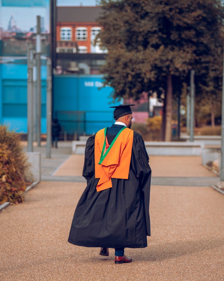 A graduate alone on the pavement in gown and cap, from behind