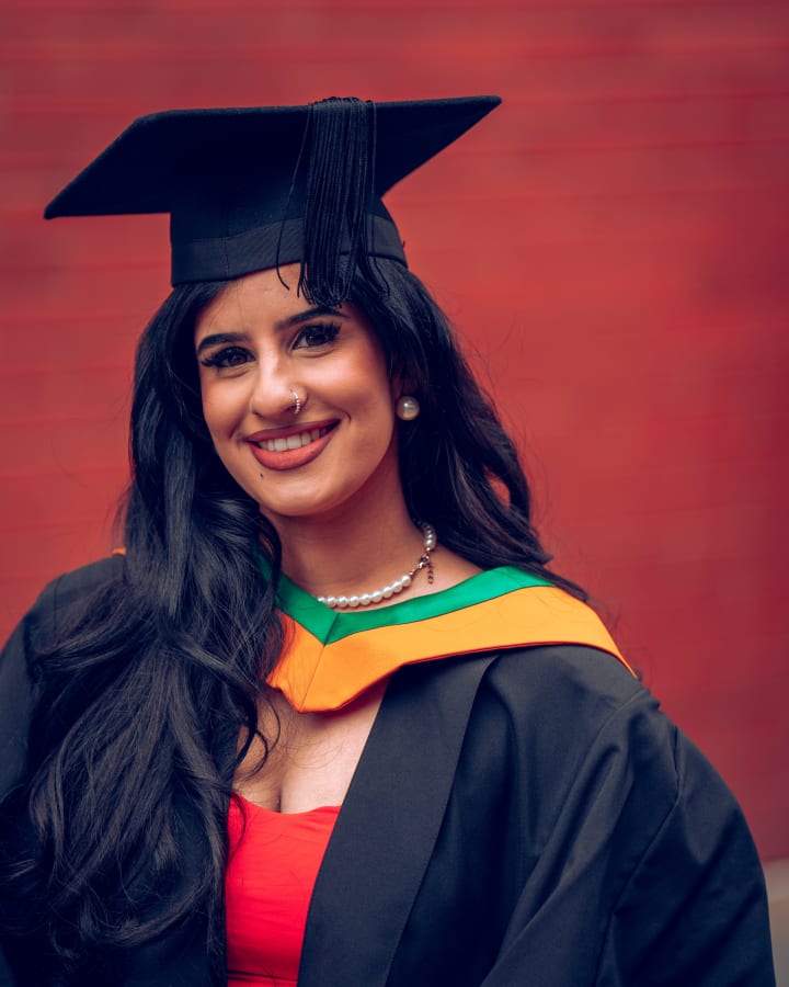 A graduate in a green and gold hood against the red wall, smiling