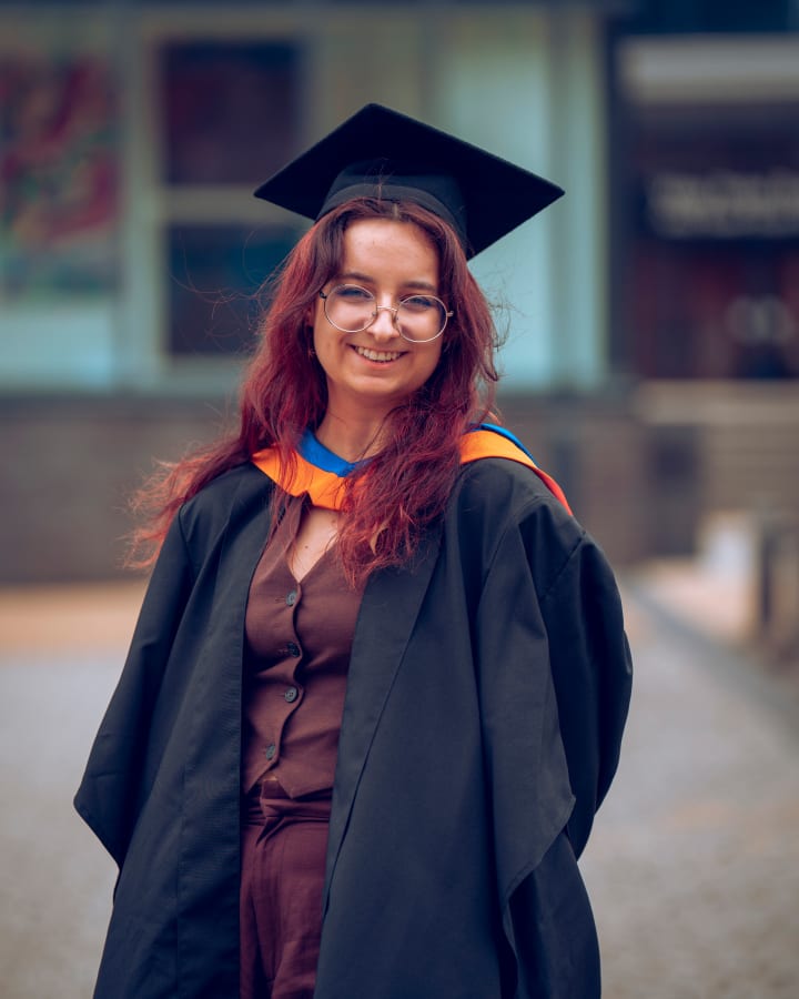 A graduate on the street, serious, the city behind her
