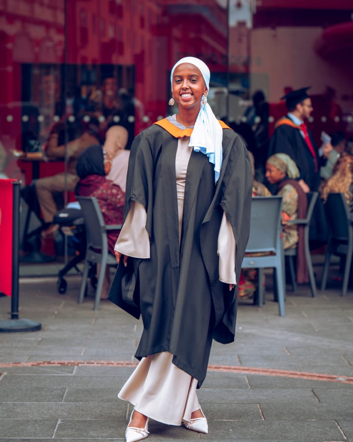 A graduate in a hijab walking towards the camera, gown open over white