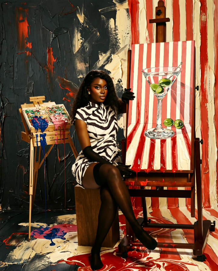 Ejiro seated on a stool in a zebra dress and opera gloves, beside an easel painted with a martini