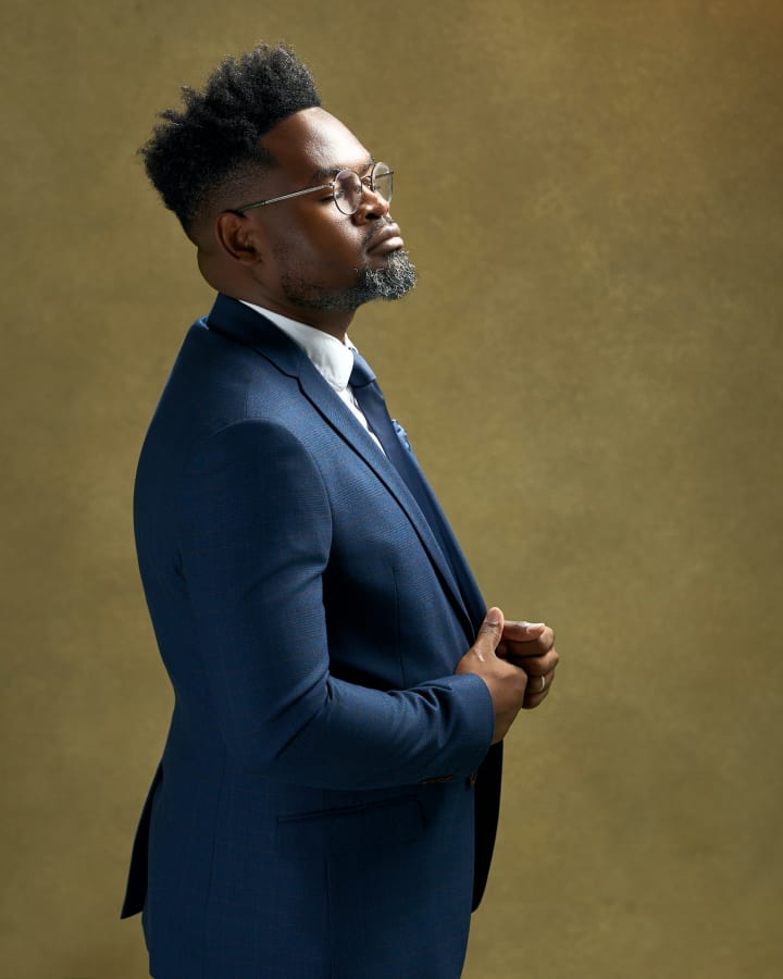 Ndirpaya in profile in a blue suit and round glasses, eyes closed, hands clasped, against an olive backdrop