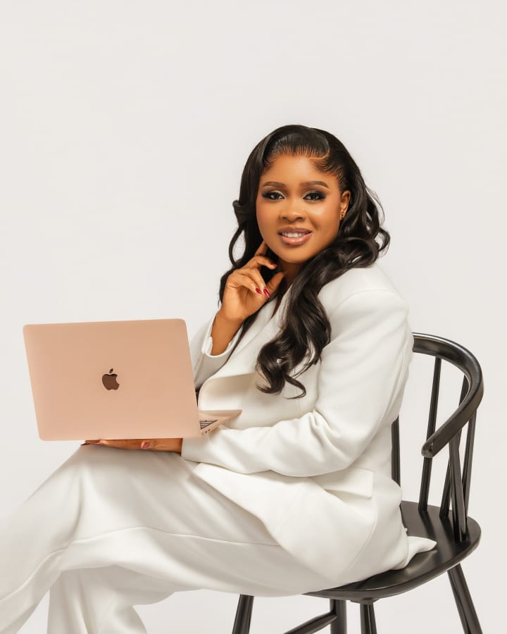 On a black Windsor chair with her laptop, a hand at her chin