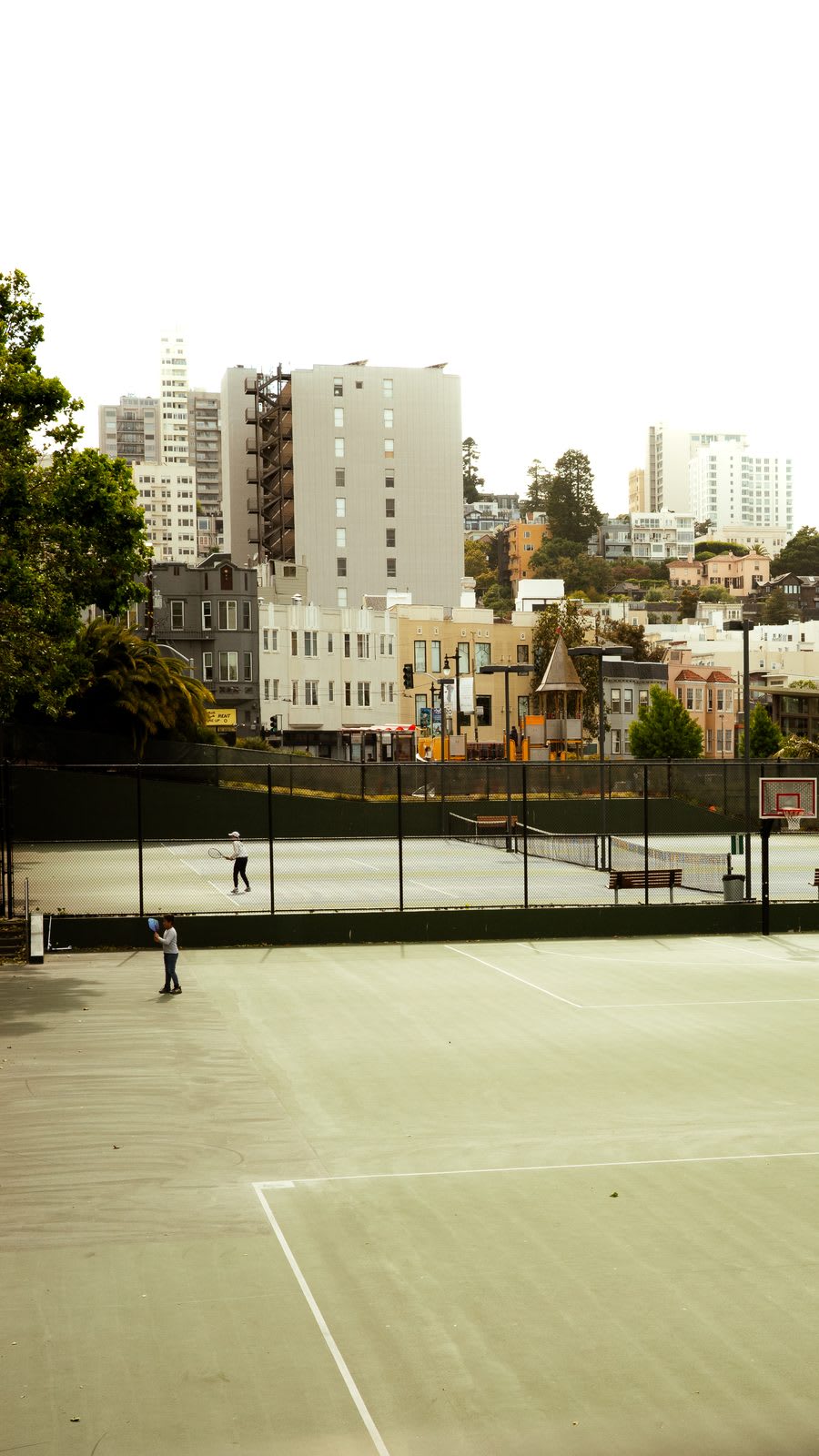 Two figures in white playing tennis on an open court, the pastel Edwardian houses of the city rising behind them under overcast sky.